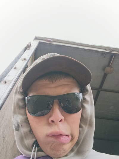 The image shows a close-up, selfie-style portrait of a person wearing sunglasses and a hooded sweatshirt. The person's mouth is slightly open, with their tongue sticking out. They are also wearing a baseball cap. The background appears to be the interior of a truck or trailer, with metal framing visible. The lighting is overcast, suggesting it is daytime. No specific details of Emel'yanovskaya, Russia, are visible in the image.