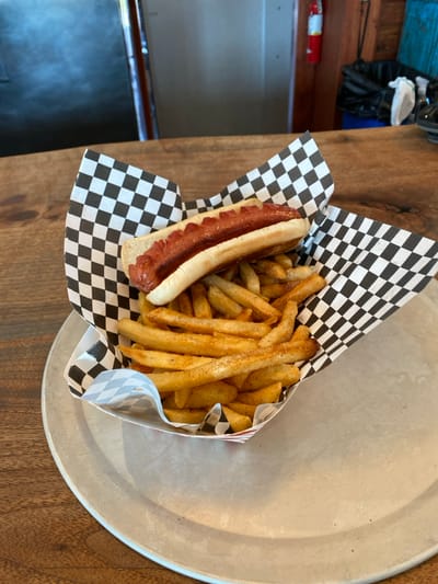 A hot dog and a generous portion of french fries are displayed in a paper boat container featuring a black and white checkerboard pattern on its rim, resting on a round, silver-colored metal tray. The tray is situated on a medium brown wooden surface, likely a table or countertop with visible grain. The hot dog is a single, scored frankfurter placed within a plain white bun. The french fries are golden brown, medium cut, and appear seasoned with dark specks. The background on the left side of the image reveals a blurred, reflective metallic surface. To the right, a red fire extinguisher is mounted on a wall, and a black trash bag is visible within a bin. A turquoise decorative object is also present on the wall behind the bin. The wall itself exhibits wooden paneling. The scene is located in Accord, United States, within an indoor commercial or dining establishment. No people or direct actions are visible.