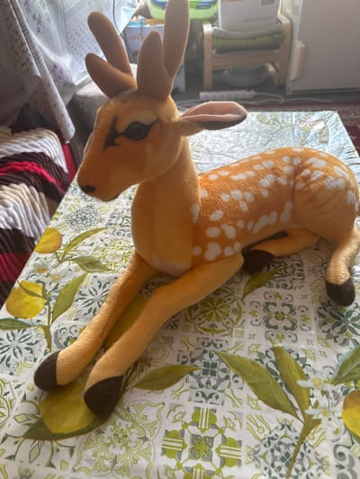 A plush toy deer with orange fur and white spots is lying on a tablecloth with a pattern of green leaves and white flowers. The deer is in focus, and its body is angled towards the right. Its head is turned to the left, with large black eyes and small brown antlers. The background is blurred, showing furniture and a window. The image appears to be taken indoors, likely in a residential setting. There is no indication of the location being Hradec Králové, Czechia.