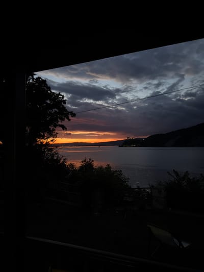 This image captures a sunset over the Danube River near Golubac Fortress, in Golubac Municipality, Serbia. The sky is filled with dramatic, dark clouds tinged with orange and purple from the setting sun, which is visible as a bright band on the horizon. A silhouette of a dense tree line and foliage frames the left side of the scene. Across the river, the dark silhouette of a hill rises, topped with the prominent Golubac Fortress, illuminated by a few lights. The water of the Danube is dark and reflects the sky's colors. In the foreground, the scene is predominantly dark, suggesting a shadowed viewpoint, possibly from a balcony or indoor seating area. There are indistinct outlines of outdoor furniture and railings in the foreground, obscured by the low light. No people are visible.