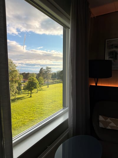 The image depicts a view from an indoor window overlooking a vibrant green lawn dotted with trees, a white building, and distant hills under a partly cloudy sky. On the right side of the frame, an interior scene includes a dark armchair with a patterned cushion, a floor lamp, and a framed artwork on the wall, illuminated by an orange backlight. The window itself shows a smudge on the glass. Based on the characteristic landscape and architecture, this view is likely from a hotel or residence located in or near Östersund, Sweden, possibly with a perspective towards the Frösön island or Lake Storsjön.