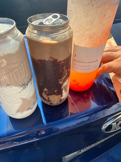 The image shows three drinks on the hood of a blue car. The drink on the left is in a clear jar with a white lid, filled with a layered drink that appears to be a milkshake or smoothie. The middle drink is in a clear can with a silver pull-tab lid, also containing a layered drink, possibly chocolate and cream. The drink on the right is in a clear plastic cup with a white label that reads "J Strawberry Lemonade Large - Iced, (Lemonade) 5:26 PM Jun 27, 2026 #89". A hand is visible reaching for this drink. The background is out of focus but suggests an outdoor setting with sunlight. The car appears to be parked, and the chrome emblem of "K AUTOMOTIVE GROUP" is visible on the hood. The image was taken in Glenview, United States of America.