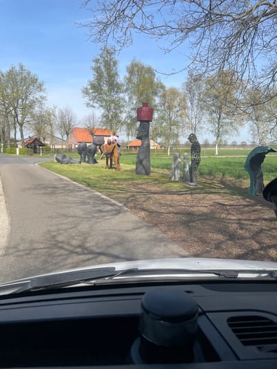 The image is taken from inside a vehicle, looking out at an outdoor scene. Alongside a road, there is a sculpture park featuring various modern art sculptures, including figures of people, animals, and abstract shapes. In the background, there are traditional European-style buildings with red-tiled roofs, indicating a rural or semi-rural setting. The sky is blue with sparse clouds, and trees with budding leaves are visible.

Based on visual markers, this is most likely the sculpture park alongside the Beeldig Geel sculpture route in Geel, Belgium.