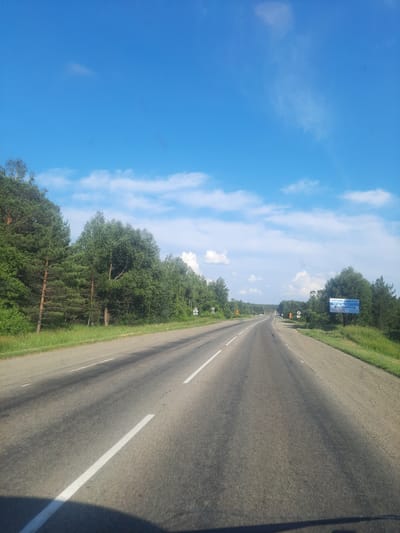 The image shows a road in the city of Баюновские Ключи, Russia. The road is asphalt, with white lane markings. On the left side of the road is a grassy area with trees, and on the right side is a sign in Russian that reads "БУРЕНИЕ СВАЛИН" and "ВОСТОЧБУРВОД". The sky is blue with some white clouds. The scene appears to be a sunny day.