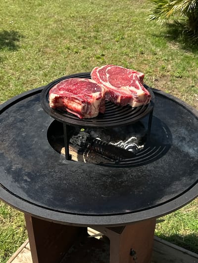 Two raw, red steaks sit on a black metal grill over smoldering wood embers. The grill is part of a larger, circular metal outdoor cooking device with a flat, table-like surface. The setting is outdoors, on a lawn with green grass and some scattered foliage. The scene appears to be in daylight. La Couarde-sur-Mer, France is not visibly identifiable in the image.