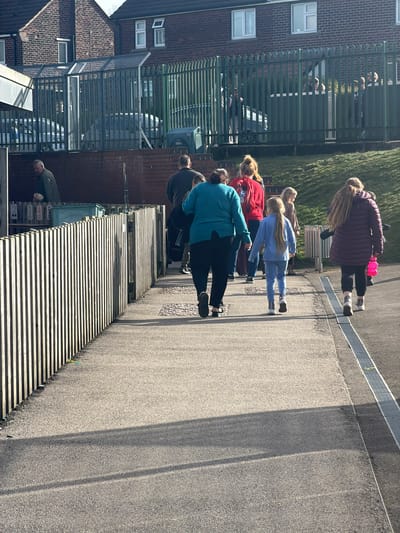 The image depicts an outdoor scene in Rotherham, United Kingdom, on a sunny day. A group of approximately ten individuals, comprising adults and children, walk away from the viewer on an asphalt pathway. In the foreground, a person in a teal top and black trousers walks centrally, accompanied by a young girl in a light blue tracksuit directly in front of them, and another young girl with long blonde hair in a purple puffer jacket holding a pink water bottle to their right. Another adult in a red hooded top is partially visible to the left. To the left of the pathway, a light-colored wooden slat fence runs parallel, behind which a low brick wall supports a series of wooden bins or storage units, some numbered '2', '3', '4', and a teal container. An adult male in a dark jacket is visible behind the wooden fence, attending to items near these bins. In the mid-ground, a tall green metal palisade fence separates the pathway area from a paved lot containing parked vehicles. Behind this fence, several additional individuals, including adults and children, are visible. To the far left, a caged structure with a translucent roof is present. In the background, two multi-story red-brick buildings with white-framed windows are visible under a clear sky. Long shadows are cast across the pathway from the left, indicating the sun's position. A metal drainage grate runs along the right edge of the pathway.