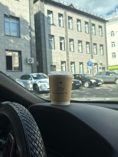 The image shows a coffee cup with the text "BAKEHOUSE" printed on it, placed on the dashboard of a car. The cup is brown and has a clear plastic lid. The background, visible through the car's windshield, depicts a building with multiple windows and an air conditioning unit. In the distance, several cars are parked on the street. The scene appears to be set in a city, likely Mahilioŭ, Belarus, during the daytime. The lighting suggests it's a sunny day, with shadows cast on the building. The overall impression is that of a casual moment, perhaps a commute or a brief stop.