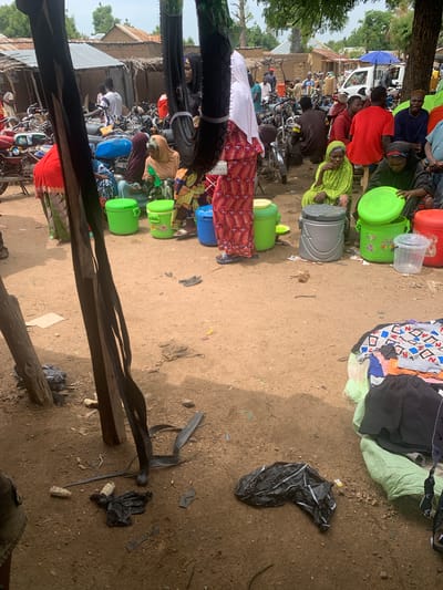 This media file depicts a bustling outdoor market scene in Nigeria, likely in the morning or early afternoon given the bright sunlight casting distinct shadows. The ground is a mixture of sandy soil and some scattered debris, suggesting a rural or semi-urban environment.
The primary subjects are a diverse group of people engaged in various activities within the market. Many individuals are dressed in colorful traditional clothing, with women often wearing headscarves and long dresses. Some people are seated, possibly vendors or shoppers taking a break, while others appear to be actively browsing or interacting. A significant number of motorcycles are parked closely together, indicating a common mode of transportation for market-goers and suggesting the market is a central hub.
A variety of objects are visible, predominantly containers. Large green plastic buckets and containers are scattered throughout the scene, some appearing new and others used. There are also a few clear plastic containers and a larger, grey, lidded bin. Piles of clothing, some folded and others loosely arranged, are also present, suggesting a clothing stall or a place where personal belongings are kept. A black plastic bag is visible on the ground, along with other assorted debris.
The overall atmosphere appears dynamic and busy. While specific emotions are hard to discern from still images, the crowded nature of the market and the presence of many people suggest a lively and active community gathering. The setting is characterized by makeshift stalls and the presence of trees and rudimentary structures in the background, further reinforcing the Nigerian market context. No visible text is identifiable within the image.