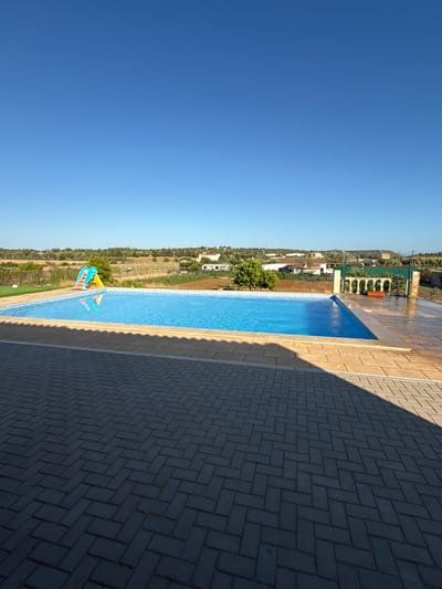 The image shows a swimming pool next to a paved area with a shadow cast by an unseen object. The pool is rectangular with blue tiles and appears to be empty of people. On the far side of the pool, there is a colorful water slide. Beyond the pool, there is a landscape with some buildings and vegetation, likely a rural or suburban area. The sky is clear and blue, suggesting a sunny day. The perspective is from a slightly elevated viewpoint looking down at the pool and surrounding area. The pavement is made of dark grey rectangular bricks laid in a herringbone pattern, with a lighter colored border next to the pool. The image is oriented vertically.