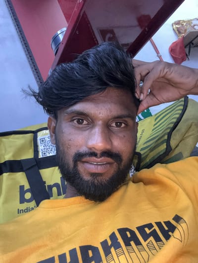 A South Asian male with a dark complexion, black hair styled upwards, and a dark beard and mustache is depicted from the chest up, looking directly at the viewer with a slight smile. He wears a yellow long-sleeved top with a dark graphic print partially visible as "THRAS...". His right hand is raised, touching the side of his head near his hairline. Behind him on the left, a yellow and black delivery bag, identified by partially visible text "b n" and "India'" (suggesting "blinkit India") and a QR code, is present. To his right, a green and yellow striped fabric item with the partial text "Minute App" is visible. The background features a reddish-maroon metal structural beam, a silver cylindrical object, a light grey wall with a slotted metal rail, and a white wall displaying a draped red fabric with gold embellishments and orange wiring. The scene is located in Palghar, India.