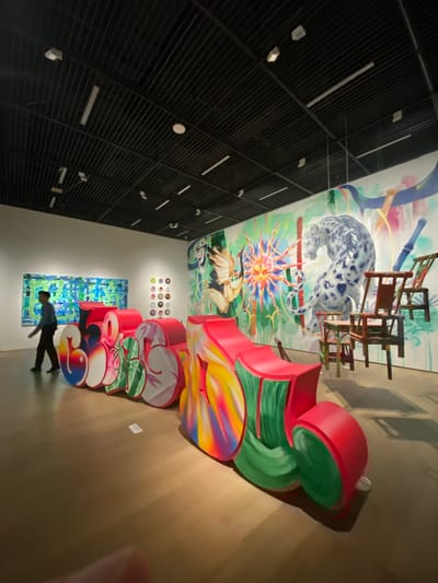This is a medium shot, eye-level, indoor photograph of an art exhibition. The exhibition space features a large, colorful mural on the back wall. The mural depicts a snow leopard, a crane, and a sun-like object amidst abstract green and blue elements. In the foreground, a large, three-dimensional sculpture of graffiti-style lettering spells out "630". The sculpture is rendered in vibrant colors, including red, pink, green, yellow, and blue, with a glossy finish. Several wooden chairs are suspended from the ceiling in front of the mural. A person, appearing to be a man in a blue shirt and dark pants, is walking from left to right in the background. The floor is a polished wooden surface, and the ceiling is dark with a grid pattern and various lights and vents. The overall impression is a modern art gallery setting. The photograph was taken in the city of Jinniu District, China.