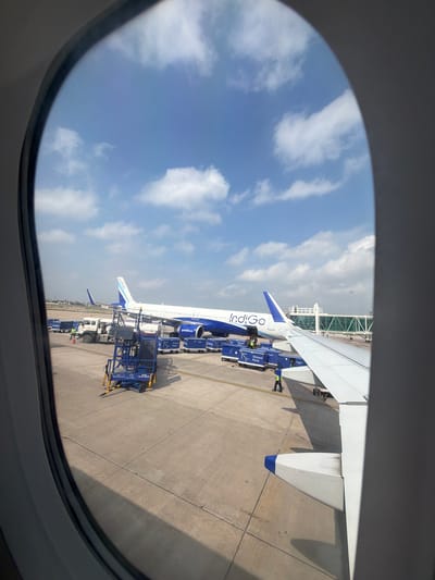 The image is a view from an airplane window looking out at an airport tarmac. In the foreground, a portion of an airplane wing is visible, extending from the right side of the frame. Beyond the wing, an Indigo Airlines airplane is parked. The airplane is predominantly white with blue accents and the Indigo logo on its tail.
On the tarmac, various airport equipment and vehicles are present. Blue cargo carts with "Indigo Ground Force" labeling are arranged near the airplane. A white truck and a blue mobile stairway are also visible. Several individuals, appearing to be airport ground crew wearing high-visibility vests, are positioned near the aircraft and equipment.
In the background, a pedestrian walkway or jet bridge connects to a building. The sky is blue with scattered white clouds. The scene appears to be a typical airport operational area under daylight conditions. No specific landmarks are visible to pinpoint the exact location within Vikram Nagar, India, but it is an outdoor airport environment.