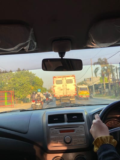 The image is taken from inside a car, looking forward through the windshield. The car appears to be in traffic, with several other vehicles visible on the road. In the foreground, a person's hand is visible on the steering wheel, wearing a yellow ruffled cuff. The dashboard of the car, including the radio and air vents, is also visible.

Outside the car, the scene is a street with moderate traffic. Two motorcycles are in the lane ahead, with riders wearing helmets. A large white truck is in the lane next to them, and a yellow truck is further ahead. Trees and buildings line the right side of the road. The sun appears to be low in the sky, casting a warm light on the scene, suggesting it's either early morning or late afternoon.

The image is identified as being taken in Tenggeles, Indonesia.