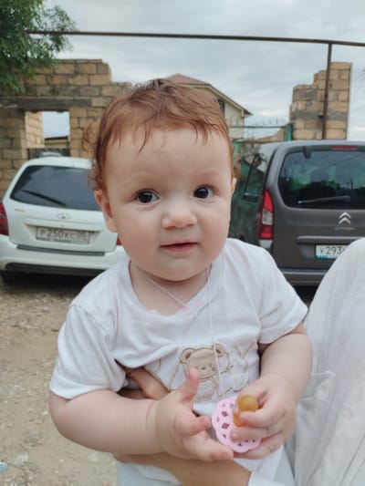 A close-up shot focuses on the face of a baby with light skin and reddish-brown hair, looking directly at the camera. The baby is wearing a white t-shirt with a graphic of a teddy bear. The baby's hands are visible, with one hand holding a pink pacifier. In the background, a white car with a license plate starting with "P 250KC" is parked, and a grey car with a license plate starting with "Y 293" is also visible. A partially constructed building made of beige bricks and some greenery are present in the background, suggesting an outdoor setting. The scene appears to be in Darvag, Russia, based on the context provided.