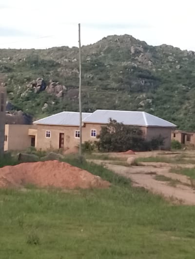 In the city of Barakin Acha, Nigeria, a long, gray, thin pole stands in the center of the frame, bisecting a light brown, single-story building with a silver, sloped roof. The building has several dark-framed windows and dark-colored doors. To the left of the main structure, another section of the building with a visible window and a dark door is present. Behind the building, a lush green hill dotted with large rocks and sparse vegetation rises. In the foreground, a pile of reddish-brown dirt is visible, with green grass surrounding it. The ground in front of the building appears to be a dirt path.