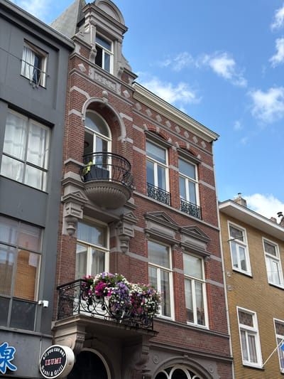 The image depicts a close-up view of the facades of several buildings in Roermond, Netherlands, under a bright blue sky with scattered white clouds. The central building is constructed of red brick with white trim, featuring ornate architectural details such as a decorative cornice, diamond-shaped accents, and a arched window with a black wrought-iron balcony above it. This balcony, like the one lower down on the same building, is adorned with a profusion of colorful flowers in shades of pink, purple, and white.

To the left of the brick building is a narrower, modern building with a dark gray facade and large, multi-paned windows. Some of these windows have white curtains, and one has a bamboo blind. A small white security camera is mounted on this facade. Below the windows of the gray building, a portion of a sign for "all you can eat IZUMI Sushi & Grill" is visible, featuring blue and white lettering on a black background.

To the right of the brick building is a building with a light yellow, horizontally ribbed facade. This building has several white-framed windows, some of which are smaller and appear to have decorative colored glass panes. A white flagpole extends from this building, though the flag it carries is only partially visible and appears to be blue and white. The overall impression is of an urban street scene with a mix of architectural styles, from historic brickwork to more contemporary designs.
