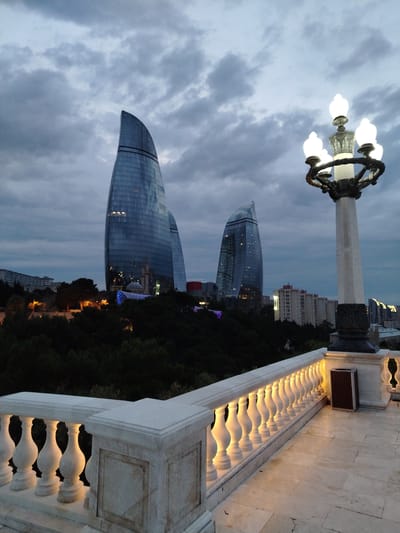 A twilight view of the Flame Towers in Baku, Azerbaijan, is captured from an elevated vantage point. The three skyscrapers, designed to resemble flames, dominate the skyline with their sleek, glass facades reflecting the dusky sky. The left-most tower is the tallest and most prominent. Below the towers, a dark, tree-covered hillside slopes downwards. Scattered lights from buildings and structures are visible on the hillside, including a dome and a minaret, suggesting a mosque or other significant edifice. A modern, curved structure with blue lighting is also visible amidst the trees.

In the foreground, a white marble balustrade with ornate balusters runs along the edge of an observation area or balcony. The balustrade is illuminated from below by warm, yellow lights, casting a glow on the white stone and the paved stone floor. A dark, rectangular trash receptacle is positioned near the balustrade. To the right, a decorative lamppost with multiple glowing white lights stands tall against the cloudy sky. The sky itself is filled with dramatic, dark grey clouds, indicative of an overcast evening. The overall mood is serene and somewhat dramatic, illuminated by the artificial lights against the natural twilight. The scene suggests a public viewing area, likely a park or promenade, offering a panoramic view of the city's modern architecture. No people are visible in the frame.