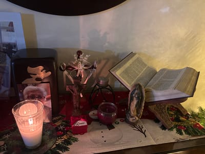 A small altar displays religious and sentimental items. A lighted candle sits beside a photo of an elderly woman. A brown urn with a dove symbol stands nearby. A cross with floral embellishments is positioned next to a small red gift box labeled "Rouss." A large open Bible rests on a wooden stand, accompanied by a statue of the Virgin Mary. The scene is arranged on a table runner with a festive Christmas design.
