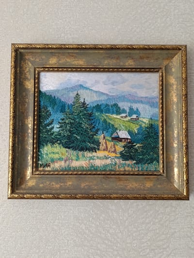 The image depicts a framed embroidered artwork of a landscape scene. The artwork is framed with an ornate gold and green frame and shows a rural scene with rolling hills, evergreen trees, a field with haystacks, and several houses. A mountain range appears in the background under a cloudy sky. The embroidery is signed "MIA" in the bottom left corner. The image is centered on the artwork, which is hanging on a light-colored textured wall. The scene does not depict the city of Barvinok, Ukraine.