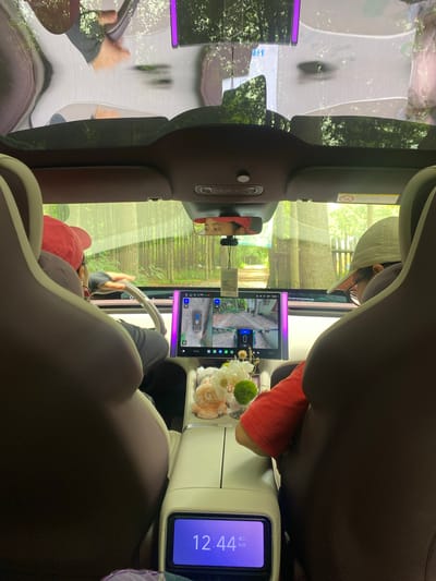 The image captures the interior of a car, looking forward from the back seat. Two individuals are visible. In the driver's seat, a person wearing a red baseball cap and glasses is holding the steering wheel. In the passenger seat, a person wearing a beige baseball cap and glasses is also looking forward. Both appear to be in motion, as suggested by the reflections on the windshield and sunroof, which show trees and foliage. The car's dashboard features a large touchscreen display showing a rearview camera feed, indicating the car is likely in operation. A digital clock on a lower console screen displays "12:44" and "6/23", suggesting the time and date. The overall environment appears to be a natural, possibly rural or suburban setting with dense trees visible through the windows. There are no overt indications of the city of Dujiangyan City, China.
