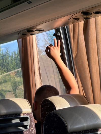 The image shows the interior of a bus, likely in Kiosevtsi, Bulgaria. Sunlight streams through the window, illuminating the arm and hand of a person in the foreground. The person's arm is extended upwards, with their index and middle fingers raised in a "V" or peace sign. The person's head and the back of their neck are visible, with short, dark hair. Two beige and patterned bus seats are in the foreground, with the one on the left partially obscured by shadow. Brown curtains are drawn on the right side of the bus. Outside the window, trees and a bright blue sky are visible. The bus ceiling features air vents and lighting fixtures.