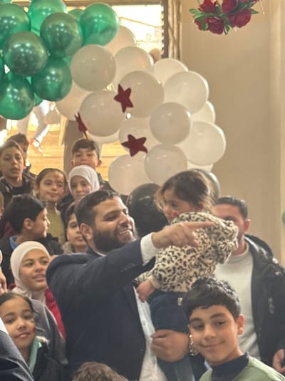 An indoor scene in Kafr Batna, Syria, depicts a group of individuals, predominantly children, gathered under a balloon arch. A bearded adult male, positioned centrally, is holding a small child wearing a leopard-print jacket. The man is smiling and pointing with his right hand towards an unseen area to the viewer's right. The child in his arms is also looking in that direction, with a faint red mark on her left cheek. Several other children of varying ages are visible around them, some smiling, and some wearing headscarves. In the background, an arrangement of green and white balloons forms an arc, with some white balloons featuring red star decals. A red floral decoration is partially visible in the upper right quadrant of the image. Other adults are partially visible in the background. The lighting suggests an interior space with artificial illumination.
