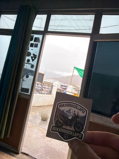 The image, taken indoors looking outwards, shows a hand holding a sticker with the text "MAXXOOOOCOLLAPSE" and an image of a motorcycle with mountains in the background. Through the window, a glimpse of the town of Nyoma, India is visible. The scene includes a green flag fluttering in the breeze and mountains in the distance under an overcast sky. Part of a building with a white railing is also visible. The window frame and a teal curtain are prominent in the foreground, along with some geometric shapes affixed to the windowpane.