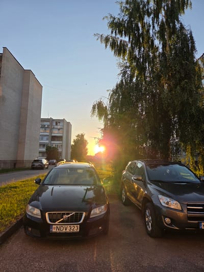 This is a photograph taken in Širvintos, Lithuania, likely during the late afternoon or early evening as indicated by the low angle of the sun and the warm, golden light illuminating the scene. The sky is clear and blue, suggesting fair weather.

The image features a street scene with several cars parked along a paved road, bordered by a grassy area and trees. In the foreground, a black Volvo sedan is prominently displayed, its front end facing the viewer. The license plate reads "NDV 323" with "LT" visible, confirming the Lithuanian context. To the left of the Volvo, the front of a dark-colored SUV is visible. Behind these vehicles, apartment buildings are visible, one with a beige brick facade and another taller, lighter-colored building with multiple windows.

The sun is low on the horizon, casting a bright lens flare and casting long shadows. The trees, with their lush green foliage, are illuminated by the setting sun, creating a warm and tranquil atmosphere. The overall impression is one of a quiet residential area at the end of a pleasant day.