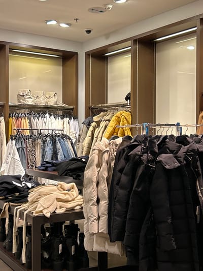 This media file captures the interior of a well-lit clothing store, presumably located in Chalkida, Greece. The perspective is slightly angled, looking into a section of the store filled with merchandise.
The dominant subjects are various articles of clothing displayed on multiple racks and shelves. On the upper racks, a prominent collection of puffer jackets or winter coats is visible, primarily in black, beige/cream, and a bright yellow. Below these, further racks hold a diverse range of garments including denim jeans, knit sweaters in light colors, and patterned tops. Some items appear neatly folded on shelves. Towards the bottom right, several pairs of black boots or shoes are neatly arranged on a dark display unit. On the far left, a shelf unit integrated into the wall, made of dark wood, displays several light-colored handbags or backpacks, illuminated by linear LED strip lights.
The setting appears modern and organized. The store features dark wooden shelving units that frame white wall sections, also illuminated by integrated LED strips. The ceiling, visible on the left side, is white and includes recessed spotlights, a round white sensor or smoke detector, and a small black security camera. The overall lighting is bright and artificial, designed to highlight the merchandise.
There are no people visible in the image, suggesting the photo might have been taken during non-operating hours or a quiet moment. No specific activity is captured beyond the static display of goods. The time of day cannot be determined from the indoor lighting. There is no discernible text on any products, signs, or labels within the frame. The scene conveys a clean, retail environment ready for customers.