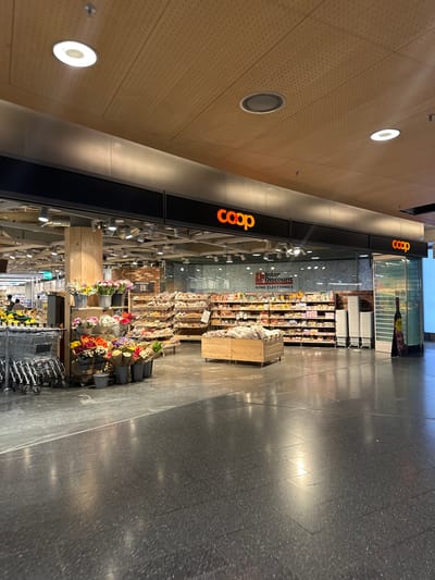 The image shows the interior of a Coop supermarket located in Kloten, Switzerland. The Coop logo is prominently displayed in orange above the store entrance. The store is well-lit with circular ceiling lights and spotlights illuminating the merchandise. The floor is dark gray with a speckled texture. Aisles are stocked with various items, including groceries, flowers, and electronics. Shopping carts are visible near the entrance. A sign for "Interdiscount Home Electronics" is visible in the store. The environment appears clean and modern. No individuals are easily identifiable.