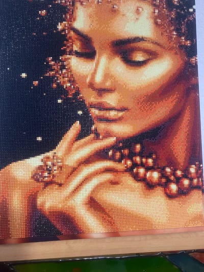 The image displays a close-up of a diamond painting depicting a woman's face. The artwork is rendered in warm tones of orange, gold, and brown, with a dark background. The woman's eyes are closed, and her head is tilted down. She is adorned with jewelry, including a large ring on her hand and a beaded necklace. The diamond painting is framed, and the frame appears to be made of wood. The background is dark, with scattered specks of light, possibly representing stars or glitter. The image does not contain any information that would allow for the determination of its location in Achinsk, Russia.