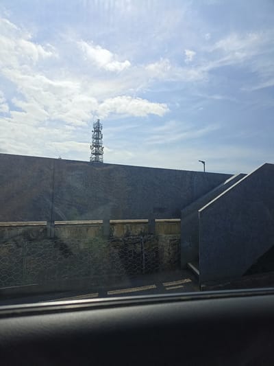 A cell tower stands tall against a bright, cloudy sky. In the foreground, a dark, textured surface, possibly a car interior, occupies the bottom portion of the frame. Beyond this, a grey, textured wall stretches horizontally, with a concrete structure featuring an angled upper section to the right. Below the wall, a wire mesh fence is visible, with a glimpse of a paved area and some greenery beyond. The scene appears to be an outdoor urban or industrial setting.