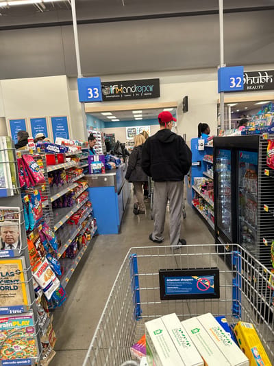 This image captures the interior of a brightly lit retail store, likely a supermarket or general merchandise store, focusing on a checkout area. The scene is presented sideways.

Several people are visible in the store. In the foreground, a person wearing a red cap, a black jacket, and gray sweatpants stands facing away from the camera, appearing to be in a checkout line. Another person in a black shirt is partially visible behind them. Further down the aisle, a person with blonde hair is seen behind a checkout counter.

The checkout lanes are numbered, with "32" associated with a "BEAUTY" section and "33" marking an "ifixandrepair" service that handles "Smartphone | Tablet | PC | Laptop." Adjacent to the checkout, a refrigerated display unit holds various bottled beverages, including Pepsi.

The foreground also shows part of a shopping cart, which contains boxes of "Beech-Nut" baby food (with descriptions like "9 fruit, banana & strawberry" and "favorite flavors") and an "Attention shoppers" sign featuring a "no entry" symbol. Surrounding the checkout area are shelves and racks stocked with impulse purchase items such as colorful bags of snacks (chips, candy) and various magazines, including titles like "Brain PLAY" and "First exploring Great Ancient World." Other signs visible include "Search for an item" and "Follow the map."

The overall atmosphere suggests a typical retail environment with customers waiting to make purchases.