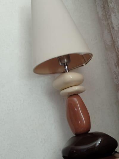 A close-up, vertical shot focuses on a decorative table lamp with a conical, cream-colored lampshade. The lampshade is slightly tilted, revealing the metallic socket and a portion of the bulb inside. Below the shade, the lamp's base is constructed from stacked, smooth, organic shapes. The top two elements are pale, ovular pieces, reminiscent of polished stones or pebbles. Beneath these is a glossy, reddish-brown, elongated oval piece. The very bottom of the lamp's base is a large, dark brown, flattened oval shape, also with a glossy finish. The lamp is positioned against a textured, light-colored wall, possibly wallpaper. To the right of the lamp, a sheer, patterned curtain is visible, suggesting the lamp is near a window. The lighting appears to be artificial, possibly from the lamp itself or an ambient indoor light. There is no visible indication of the time of day, weather, or any specific emotions. The scene is likely an indoor setting in a residential or commercial space in Yarovoye, Russia. Faint, partial text is visible on the metallic part of the lamp socket, appearing to be electrical specifications like "220-240V 50/60Hz".