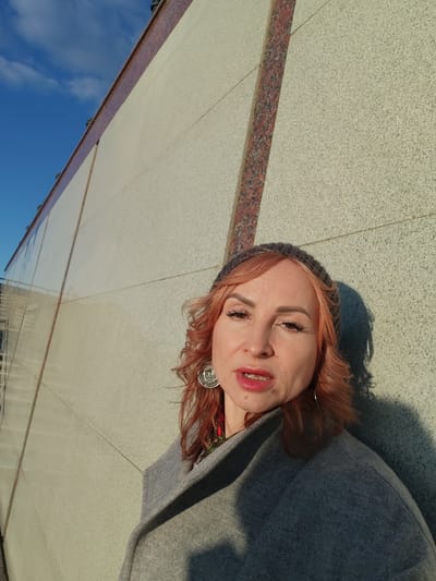 A woman stands next to a wall, likely outdoors during the day. The wall is a light-colored material with sections divided by vertical strips of red marble. She is wearing a gray coat, a knit cap, and a large earring. The sky is visible in the background with blue and some clouds. The location is in the city of Люк, Russia.