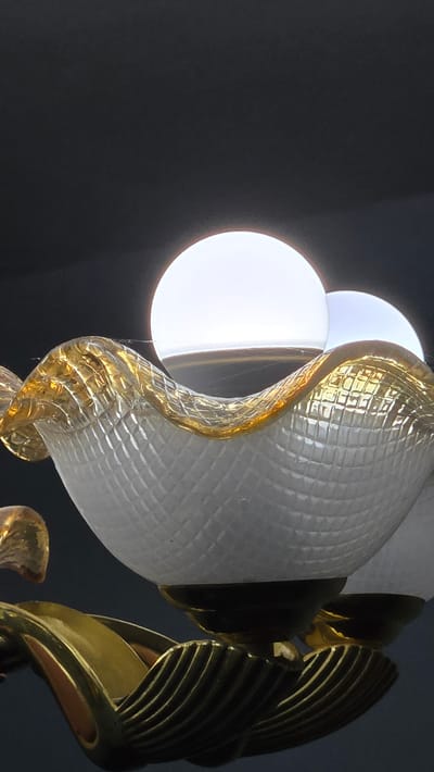 A close-up, vertical, low-angle shot captures a decorative light fixture against a dark background. The fixture features a frosted glass shade with a textured, diamond-like pattern and a golden, wavy rim. Two luminous white light bulbs are visible within the shade, one prominently in the foreground and another partially visible behind it. Delicate spiderwebs are visible on the glass shade, suggesting the fixture may be in a dimly lit or infrequently used area. The base of the fixture appears to be ornate brass, with curved, fluted elements. The location in Kaka, Turkmenistan is not discernible from the image itself.