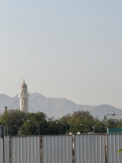 A landscape shot showing a minaret nestled amid verdant trees, framed by mountains in the background. The structure is predominantly white with a distinctive dome. In the foreground, metallic horizontal panels, likely part of an architectural feature, add texture to the composition. Streetlights dot the scene, lining the road beside the trees. The sky is a soft, muted hue, contributing to a serene, almost ethereal atmosphere.