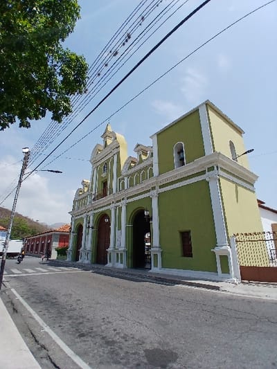 An outdoor, wide-angle street-level photograph depicts the facade of a large, historically-styled building, identified as a church, in San Joaquín, Venezuela, under a clear, bright blue sky with minimal cloud cover. The primary structure is painted an olive green, featuring cream-colored architectural details including prominent pilasters, cornices, and decorative elements. The facade presents at least three large arched entryways with dark wooden double doors. Above the central arch, a small balcony with a balustrade is visible. To the right, a square bell tower, also green with cream trim and an arched opening housing a bell, rises above the main structure. Further right, a cream-colored wall section extends, partially obscured by a brown metal fence with intricate patterns.
In the foreground, an asphalt road occupies the lower portion of the image, displaying cracks and dark stains. A white curb borders a narrow sidewalk on the left. A faded white pedestrian crosswalk is marked on the asphalt near the sidewalk. On the far left, a black utility pole with an attached streetlight stands. Numerous black utility lines crisscross the upper sky, some bearing small clusters of vegetation. A dense green tree canopy extends into the upper left corner, partially obscuring the sky and power lines.
In the mid-ground, beyond the utility pole and across the street from the church, a row of single-story buildings with red-tiled roofs and red-brown walls are visible. A white box truck is partially obscured behind the utility pole. A single individual on a black motorcycle is observed traveling away from the camera on the street, positioned between the white truck and the red-brown buildings. In the distant background, green, tree-covered hills or mountains rise under the clear sky. The scene is illuminated by direct sunlight, indicating daytime conditions.