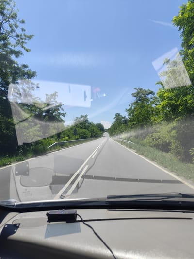 The image shows a view from inside a vehicle looking out through the windshield. The vehicle is on a paved road, which appears to be a two-lane highway, with white lines marking the lanes. The road curves gently to the left and stretches into the distance, disappearing over a slight rise. On either side of the road, there are dense green trees and vegetation, suggesting a rural or forested area. The sky is clear and bright blue with a few faint wisps of clouds or contrails. There are reflections on the windshield, showing some of the interior of the vehicle and the sky. In the foreground, on the dashboard, a small black electronic device with colored lights is visible. The image was taken in Malko Tarnovo, Bulgaria, on a sunny day.