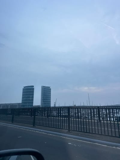The image displays a waterfront urban landscape at Chatham Maritime, adjacent to Gillingham, United Kingdom. The view is captured from within a vehicle, with the partial reflection of a side mirror visible in the lower left foreground.
In the immediate foreground, a dark grey paved surface with a thin white line is visible. A black metal railing, composed of vertical bars, spans horizontally across the middle ground, delineating the edge of a roadway or bridge.
Beyond this railing, a marina is densely packed with numerous docked vessels, predominantly sailboats, characterized by their prominent vertical masts. In the background, two tall, modern, cylindrical multi-story buildings with extensive glass exteriors, identified as the Pier Quays Towers, stand against the horizon. To the left of these towers, a lower, elongated building is discernible. A large industrial crane is positioned behind the marina, to the right of the left tower.
The sky is uniformly overcast, exhibiting a light grey-blue hue. Faint contrails are present in the upper right portion of the sky. No individuals or active interactions are observed within the frame.