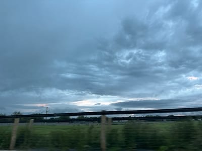 A wide-angle, eye-level shot captures a cloudy sky over a rural landscape. The sky is filled with a dense layer of gray and blue clouds, with hints of pink and white peeking through near the horizon. A dark, horizontal fence stretches across the middle of the frame, obscuring the view of the fields and trees below. In the distance, a utility pole with wires is visible, suggesting the presence of infrastructure in this undeveloped area. The overall impression is one of a calm, overcast day in the countryside, possibly near Rathvilly, Ireland, given the hints of vegetation and the type of fence seen.