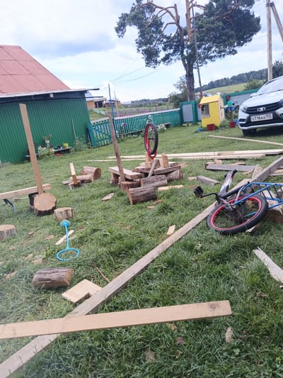 The image depicts a yard in Islambaevo, Russia, with a focus on construction materials and children's toys scattered on the grass. A green wooden fence encloses part of the yard, with a red-roofed building visible behind it. Several wooden planks and logs are present, suggesting ongoing construction or repair work. A bicycle with red rims lies on its side, and another bicycle wheel is propped up vertically. A blue plastic toy is also on the grass. In the background, a white car with a license plate that reads "O716AX 702" is parked, and a small yellow wooden structure stands near the fence. The scene is set against a backdrop of trees and a cloudy sky.