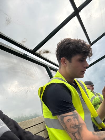 A man in a high-visibility vest is seated under a shelter with a corrugated plastic roof and walls. He has dark, curly hair and a tattoo of a man in a hat on his arm. To his right, another man wearing a similar vest and a cap is visible. They appear to be in a public area, possibly a bus stop or waiting area, with a wooden bench in the foreground. The sky is overcast, suggesting a cloudy day. The location is not identifiable as Boston, United Kingdom.