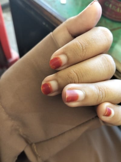 A hand rests on a beige fabric. The fingers are curved, showing nails painted with a reddish-brown hue, fading into the natural nail color at the base. The skin is light brown with visible creases on the knuckles and fingers. The texture of the fabric appears soft, slightly wrinkled, and covers part of the hand. A corner of a wooden surface with green and white writing is faintly visible in the background.