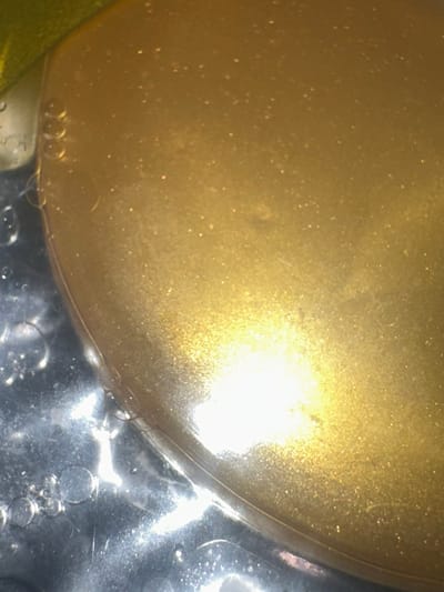 The image is a close-up, macro shot of a golden, viscous liquid, possibly oil or a cosmetic product, with fine shimmering particles suspended within it. The liquid has a smooth, glossy surface that reflects light intensely, creating a bright, hazy area in the center and lower right portion of the frame. The left side of the image shows a transparent container or surface with several small, spherical bubbles. A thin, curved line, which could be a reflection or an edge of the container, separates the golden liquid from the bubbly area. The background is out of focus and appears to be a dark surface.

There is no identifiable information or context within the image that would allow for its location in Letterkenny, Ireland, or any other specific city. The image focuses solely on the texture and reflective properties of the liquid and bubbles.