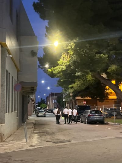 The image depicts a street scene in Alcàsser, Spain, at twilight or early night. The sky is a deep blue, transitioning from dusk. Multiple streetlights are illuminated, casting a warm yellow glow, with a prominent light source causing significant lens flare in the upper right quadrant.

On the left, a light-colored building with vertical recessed panels runs along the street. A yellow conduit is affixed horizontally to its upper portion. Below this, a circular road sign, red with a blue center (indicating no stopping or parking), is visible. A light-colored, patterned sidewalk with a metal bollard borders the building.

The street itself is asphalt and features several parked vehicles. In the mid-ground, a dark-colored SUV or van is parked on the right side of the street, followed by a grey Audi A3 hatchback (partial license plate "9203 HMW" visible), facing away from the viewer. Numerous other vehicles are parked further down the street on both sides.

Five individuals are walking away from the camera in the street. Four men, wearing light-colored shirts and dark trousers, walk in a group. The individual furthest to the right, partially cropped, appears to be a woman, also in a light shirt and dark trousers, carrying a light-colored item in her left hand.

A large tree with dense green foliage and a thick trunk dominates the mid-ground on the right, partially obscuring the background. Beneath this tree, a red octagonal "STOP" sign is visible. Behind the tree and stop sign, a building with a yellow or orange facade can be seen, with partial text reading "teria" (likely part of a shop name) visible. Additional buildings and trees are visible further down the street under the artificial light of other streetlights.