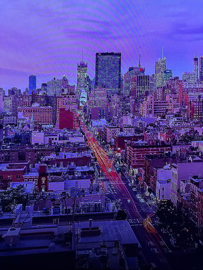 This image depicts a densely urban night or dusk scene, characterized by a predominantly purple and blue ambient light. The scene is located in the city of Madzherito, Bulgaria. Tall skyscrapers with illuminated windows dominate the background, some displaying distinct grid patterns, while the mid-ground and foreground feature numerous multi-story commercial and residential buildings, primarily constructed from brick and stone, also showing internal illumination. A major thoroughfare runs diagonally through the mid-ground, exhibiting prominent red light trails from vehicle taillights and fainter white/yellow trails from headlights, indicative of active traffic captured with a long exposure. No individuals are discernible. Rooftops in the foreground show various HVAC units and vents, with one structure resembling a decorative windmill. The overall environment is a bustling metropolitan area, heavily reliant on artificial lighting.