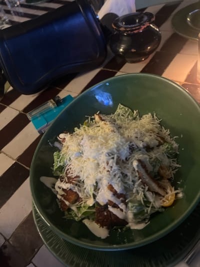 A green bowl holds a salad with greens, croutons, grated cheese, and dressing. Beside the bowl on a diagonally tiled surface are a blue purse, a small black vase, a glass, and a box. The salad appears to be a Caesar salad variant. Lighting is dim, creating shadows.