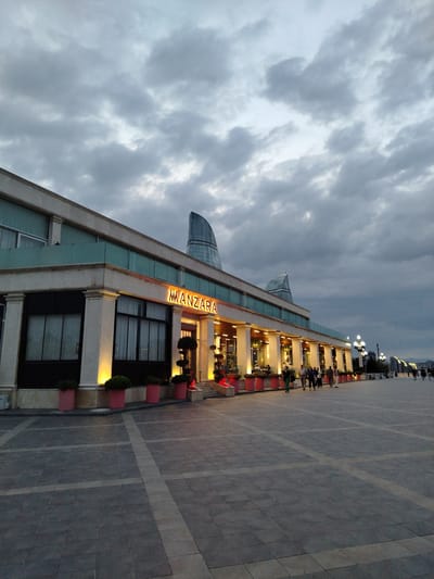 The image depicts the exterior of the Manzara restaurant, located in Sabail Raion, Azerbaijan. The restaurant is a single-story building with a white facade, featuring large windows and multiple columns supporting an overhang. A prominent orange neon sign above the entrance reads "MANZARA". The building is illuminated by warm yellow lights from within and from decorative lamps placed along the front. Several potted plants in vibrant pink containers are situated along the perimeter of the building.

In front of the restaurant, a wide, paved plaza with a patterned stone surface extends outwards. The plaza is partially populated with several individuals walking and standing. The sky above is overcast with heavy, grey clouds, suggesting an evening or pre-storm atmosphere. In the background, a modern, glass-covered skyscraper with a curved design is visible, identifying the location as a city with contemporary architecture. Along the right side of the plaza, a row of streetlights illuminates the area, and further in the distance, more buildings and lights can be seen, indicating an urban environment. The overall scene captures the ambiance of a public space in front of a dining establishment at dusk or during twilight.