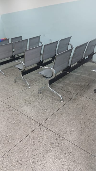 The image shows a row of empty metal chairs with perforated seats and backs, arranged along a light-colored wall. The floor is made of terrazzo tiles. The location, Barcelona, Venezuela, is not discernible from the image.